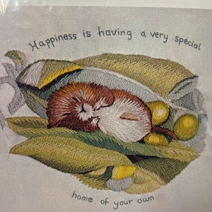May include: Embroidered artwork featuring a small brown and white mouse sleeping inside a green pea pod. The text "Happiness is having a very special home of your own" is stitched above the mouse.