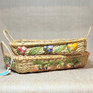 May include: Two rectangular woven baskets stacked. The top basket is decorated with colorful ribbon flowers and green leaves. The bottom basket features strawberry and leaf ribbon embellishments. Both have rope handles.