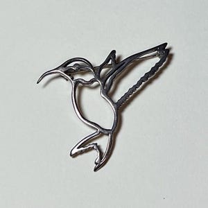 May include: A silver-colored hummingbird brooch. The brooch is a stylized outline of a hummingbird in flight, with detailed wings and a long, curved beak. The pin is attached to the back for easy wear. The brooch is a piece of jewelry.