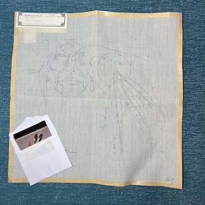 May include: Needlepoint canvas featuring a detailed outline of a Native American man's face and headdress. The canvas is beige with a yellow border. A small white envelope with a photograph is in the lower left corner. The text "Needlepoint by J" is visible.