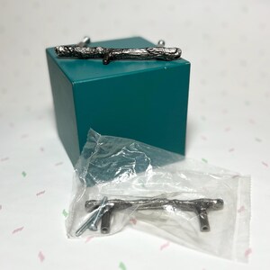 May include: Silver-toned metal cabinet pulls with a twig design. One pull is on top of a teal cube, and another is in a clear plastic bag. The pulls have a rustic, natural aesthetic, suitable for furniture or decor.