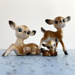 May include: Three ceramic deer figurines in varying sizes. The deer have brown and cream-colored bodies with black accents on their ears and eyes. One deer is lying down, one is standing, and one is lying down.