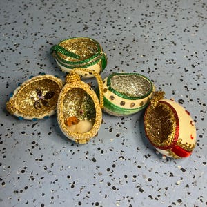 May include: Five decorative egg-shaped ornaments with open fronts, featuring cream-colored shells, gold glitter interiors, and various accents including green, red, and blue trim, small beads, and miniature internal figurines like a small brown animal.