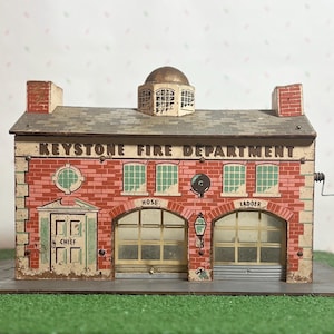 May include: A vintage tin toy fire station with a red brick facade and a white sign that reads "KEYSTONE FIRE DEPARTMENT". The toy has a working door, windows, and a bell.