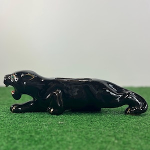May include: A black ceramic panther figurine with an open mouth and gold accents on the paws and eyes. The figurine is lying down on a green surface. The panther has a slot on its back, suggesting it is a bank.