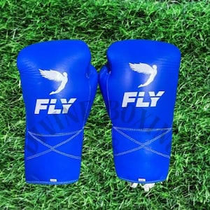Puede incluir: Un par de guantes de boxeo azules brillantes con la palabra "FLY" y un diseño de pájaro blanco. Los guantes están sobre hierba verde, lo que sugiere uso en exteriores. Guantes de boxeo para entrenamiento.