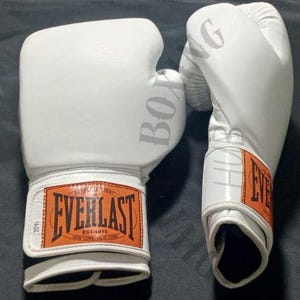 Puede incluir: Par de guantes de boxeo Everlast blancos. Los guantes tienen la palabra "BOXING" impresa en el lateral y el logotipo de Everlast en la correa de la muñeca. Las correas de la muñeca son naranjas con texto negro. Los guantes están sobre una superficie gris oscuro.
