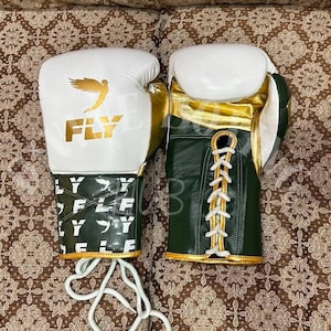 Könnte beinhalten: Ein Paar Boxhandschuhe mit einem weißen Oberteil, einem goldenen Flügel-Logo und dem Schriftzug FLY. Der Handgelenksbereich ist dunkelgrün mit einem wiederkehrenden weißen FLY-Logo-Muster. Der zweite Handschuh hat einen dunkelgrünen Körper mit weißen Schnürsenkeln.