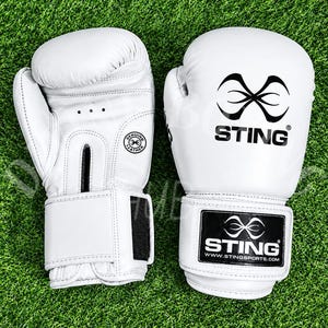 Op de afbeelding: Een paar witte bokshandschoenen met het zwarte Sting-logo op de polsband en de rug van de hand. De handschoenen bevatten een cirkelvormige stempel met de tekst Genuine Leather en drie kleine ventilatiegaatjes aan de palmzijde van de handschoen.