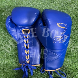 Può includere: Un paio di guanti da boxe blu reale con chiusura a lacci e bordo color cuoio. Il guanto destro presenta una grafica dorata di un uccello in volo sopra la scritta FLY. I lacci sono blu e i guanti hanno una finitura liscia e opaca.
