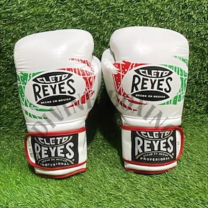 以下が含まれることがあります： 赤、緑、黒のアクセントが付いた白いボクシンググローブのペア。「CLETO REYES」のロゴ、「HECHO EN MEXICO」と「PROFESIONAL」の文字が特徴です。緑色の背景に配置されています。