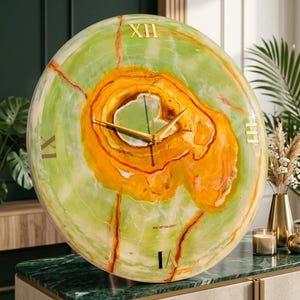 May include: A round wall clock featuring a polished green and orange stone face with natural veining. It has gold-colored metal hands and gold Roman numeral markers at the twelve, three, six, and nine o'clock positions. The text ROK ART GALLERY is printed in small black letters near the center.