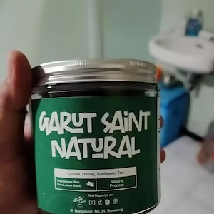 Puede incluir: Un tarro de té Garut Saint Natural, con una etiqueta verde y tapa plateada. La etiqueta presenta el texto "GARUT SAINT NATURAL" en blanco, junto con "Lychee, Honey, Sunflower Tea." El tarro es sostenido por una mano.