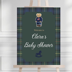 May include: A baby shower welcome sign on a wooden easel. The sign features a teddy bear wearing a sweater and jeans. The text reads "Welcome to Clara's Baby Shower" with a rocking horse illustration. The background has a green plaid pattern.