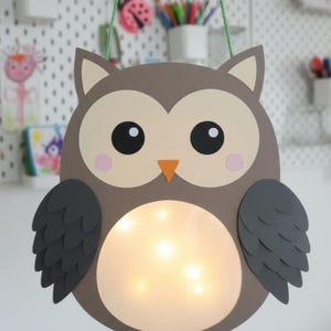 Może przedstawiać: Ręcznie robiona papierowa latarnia w kształcie sowy z ciepłym, świecącym światłem. Sowa ma brązowy korpus, beżową twarz i ciemnoszare skrzydła. Ma duże czarne oczy, różowe policzki i pomarańczowy dziób. Do zawieszenia przymocowany jest zielony sznurek.