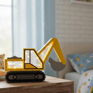 May include: A yellow and black excavator-shaped night light with a gray bucket. The night light has a black base with rolling tracks and a clear window with small lights inside. It sits on a wooden surface next to a stack of books and a teddy bear.