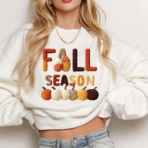 Może przedstawiać: Biała bluza z napisem "FALL SEASON" w teksturowanych, jesiennych literach i grafiką dyni. Litery są w odcieniach pomarańczu, brązu i kremu. Dynie są w różnych kolorach, w tym pomarańczowym, kremowym i brązowym.