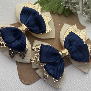 May include: Two hair bows featuring a layered design with navy blue ribbed fabric, gold glitter accents, and cream-colored ribbed fabric backing. The bows are constructed with a central navy blue knot and are displayed on a brown card backing.
