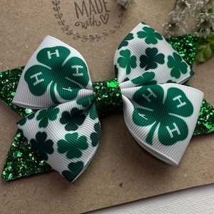 May include: A green and white bow with a four-leaf clover design and the letters "H" printed on each leaf. The bow is accented with green glitter.