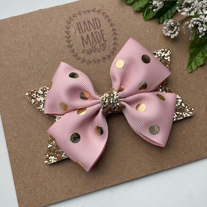 Peut inclure: Un n&oelig;ud pour cheveux rose et or &agrave; pois avec des accents de paillettes. Le n&oelig;ud est fait &agrave; la main et porte un tampon "Hand Made with Love" sur le fond.