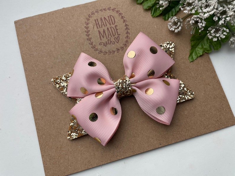 Peut inclure: Un n&oelig;ud pour cheveux rose et or &agrave; pois avec des accents de paillettes. Le n&oelig;ud est fait &agrave; la main et porte un tampon "Hand Made with Love" sur le fond.