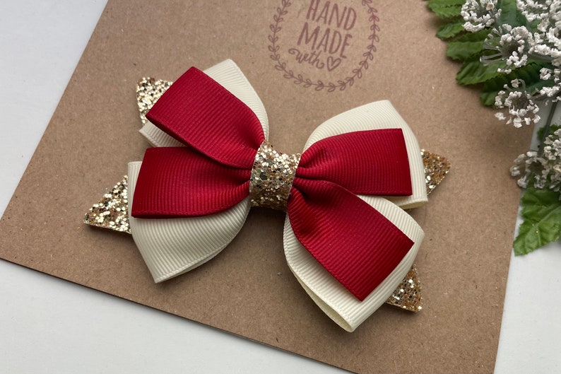 May include: A red and white bow with gold glitter accents. The bow is handmade and has a "Hand Made with Love" stamp on the background.