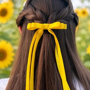 May include: A bright yellow ribbon hair bow featuring a central knot and two long, flowing tails. The bow is secured to a section of dark brown hair that is styled in a half-up braid, creating a clean and structured look for the hair accessory.