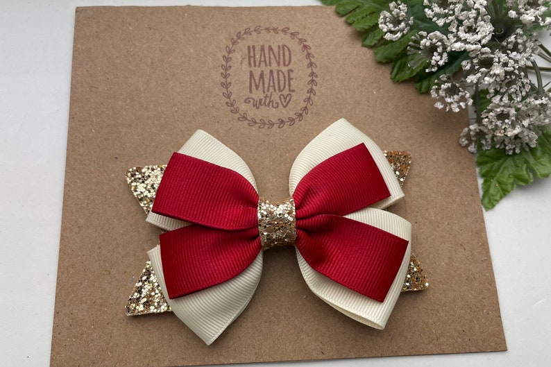 May include: A red and white bow with gold glitter accents. The bow is handmade and has a "Hand Made with &hearts;" stamp on the background.