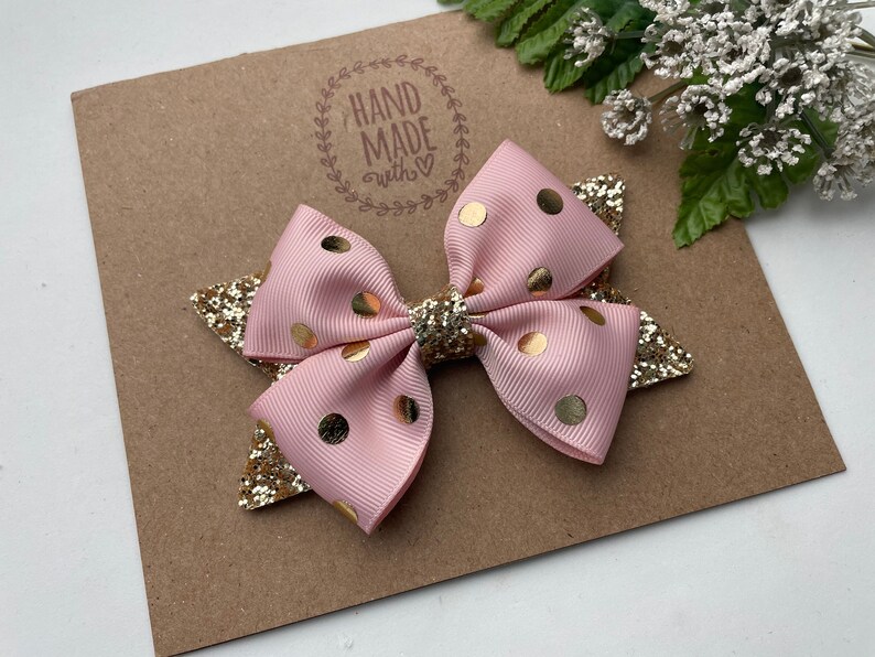 Peut inclure: Un n&oelig;ud rose et or &agrave; pois avec des accents de paillettes. Le n&oelig;ud est fait &agrave; la main et porte un tampon "Hand Made with Love" sur le fond.