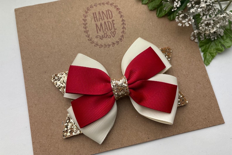 May include: A red and white ribbon bow with gold glitter accents. The bow is handmade and has a "HAND MADE with &hearts;" stamp on the background.