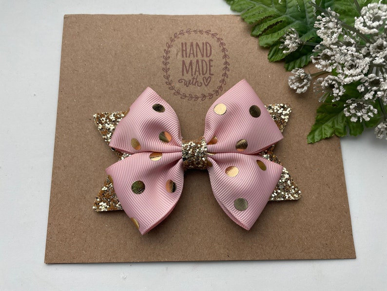 Peut inclure: Un n&oelig;ud rose et or &agrave; pois avec des accents de paillettes. Le n&oelig;ud est fait &agrave; la main et porte une &eacute;tiquette qui dit "Hand Made with love".