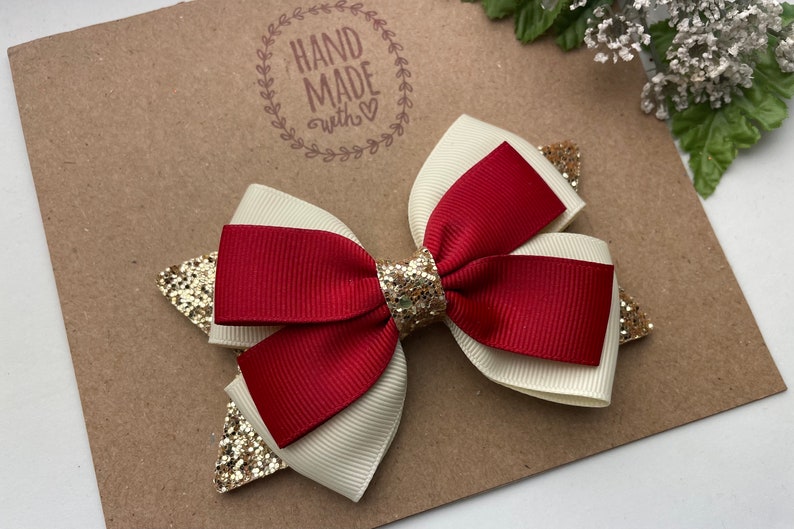 May include: A red and white bow with gold glitter accents. The bow is handmade and has a "Hand Made with Love" stamp on the background.