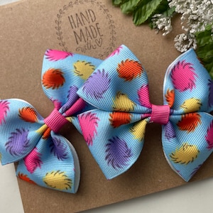 May include: Two blue hair bows with a white backing. The bows are decorated with a colorful print of fuzzy, multi-colored shapes. The bows are tied with a pink ribbon.