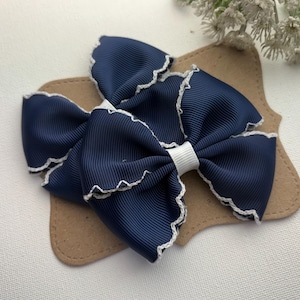 May include: A pair of dark navy blue hair bows made from ribbed fabric. Each bow features a scalloped white stitched edge and a central white ribbon band. The bows are attached to a tan-colored card backing with rounded, decorative edges.