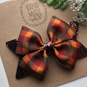 May include: A brown, orange, and black plaid hair bow with glitter accents. The bow is handmade and has a "Hand Made with Love" stamp on the background.