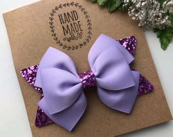Può includere: Un fiocco per capelli in tessuto a coste color viola chiaro sovrapposto a una base a forma di stella con glitter viola scuro. Il centro del fiocco è avvolto in materiale glitterato viola scuro abbinato su un cartoncino con il testo HAND MADE.