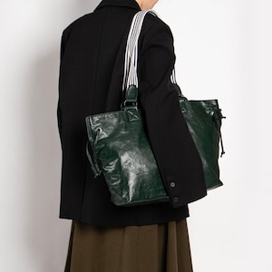 Peut inclure: Un sac fourre-tout en cuir vert foncé avec une fermeture à cordon et une bandoulière rayée. Le sac est porté à l'épaule, et la sangle a des rayures noires et blanches. La personne porte un blazer noir et une jupe marron.