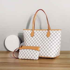 Puede incluir: Un bolso tote a cuadros blancos y grises con asas y ribetes de cuero marrón, junto con una pequeña bolsa a juego. El bolso tote tiene forma rectangular, la bolsa tiene cierre de cremallera y correa de muñeca.