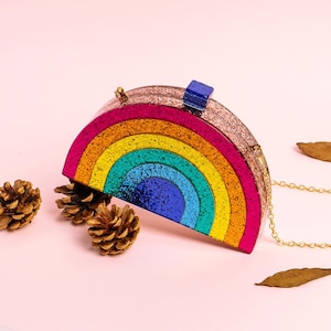 May include: A rainbow-shaped clutch with a gold chain strap. The clutch features a glittery design with bands of pink, orange, yellow, green, and blue. A small blue clasp is on top. The background is a soft pink.