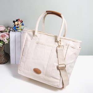 Peut inclure: Sac fourre-tout en toile crème avec une poignée en cuir marron et une bandoulière amovible. Le sac comporte plusieurs poches et un petit patch ovale en cuir. Une figurine décorative de Mickey et Minnie Mouse est visible en arrière-plan.