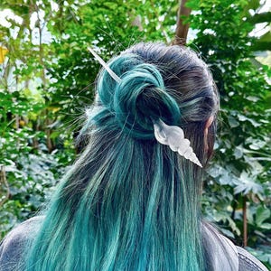 May include: A clear, seashell-shaped hair pin adorns teal and black hair styled in a bun. The hair pin is inserted through the bun and secured with a long, straight pin. The background is a lush green foliage.