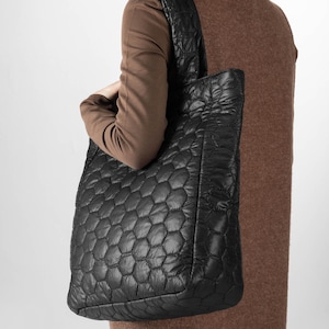 Könnte beinhalten: Schwarze, gesteppte Tragetasche mit einem geometrischen Wabenmuster. Die Tasche hat eine rechteckige Form mit einem breiten Schulterriemen. Die Tasche ist aus einem weichen, gepolsterten Material gefertigt.