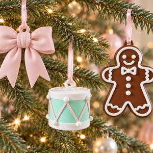 Può includere: Tre decorazioni natalizie da appendere: un fiocco rosa chiaro, un tamburo cilindrico verde menta e bianco con accenti rosa e una figura di omino di pan di zenzero marrone con dettagli in glassa bianca, appesi con nastri rosa chiaro.