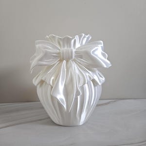 May include: A pearlescent white vase with a large, decorative bow at the top. The bow and vase have a satin-like texture, creating a soft, elegant appearance. The vase has a rounded shape with vertical ridges.