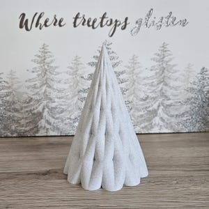 May include: A light gray fabric Christmas tree with a textured surface. The tree is in front of a white background with a forest scene and the text "Where treetops glisten".