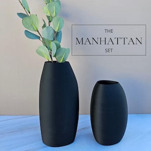 May include: Two matte black vases, one tall and one short, are displayed. The taller vase holds green and blue faux eucalyptus branches. The text "THE MANHATTAN SET" is in a black rectangle. The vases have a modern, minimalist design.