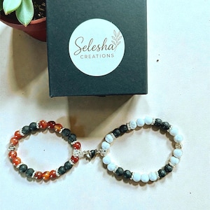 Peut inclure: Deux bracelets de perles avec fermoirs magnétiques, l'un avec des perles rouges et noires, l'autre avec des perles blanches et noires. Une boîte noire avec le texte "Selesha Creations" est en arrière-plan. Une petite plante succulente est également visible.