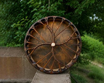 May include: A circular frame drum with a dark brown, mottled animal hide head. The wooden frame features a series of evenly spaced notches along the outer edge, with thin, dark brown leather lacing radiating from a central point to the frame.
