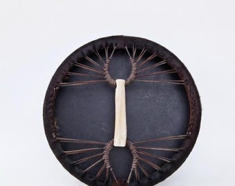 Può includere: Un tamburo rotondo di colore scuro con un manico centrale di colore chiaro. La faccia del tamburo è di un nero intenso, circondata da un materiale marrone scuro simile alla pelliccia. Corde marroni si irradiano dal centro, creando un motivo unico. Il tamburo è su sfondo bianco.