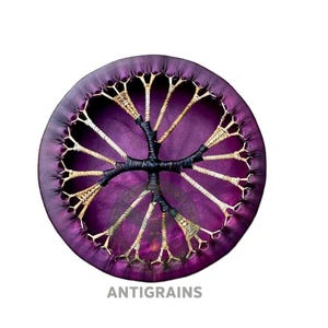 May include: A round, purple drum with a tree-like design in the center. The drumhead is a deep purple, with a stitched border. The tree design is made of black and tan woven material. The text "ANTIGRAINS" is at the bottom.